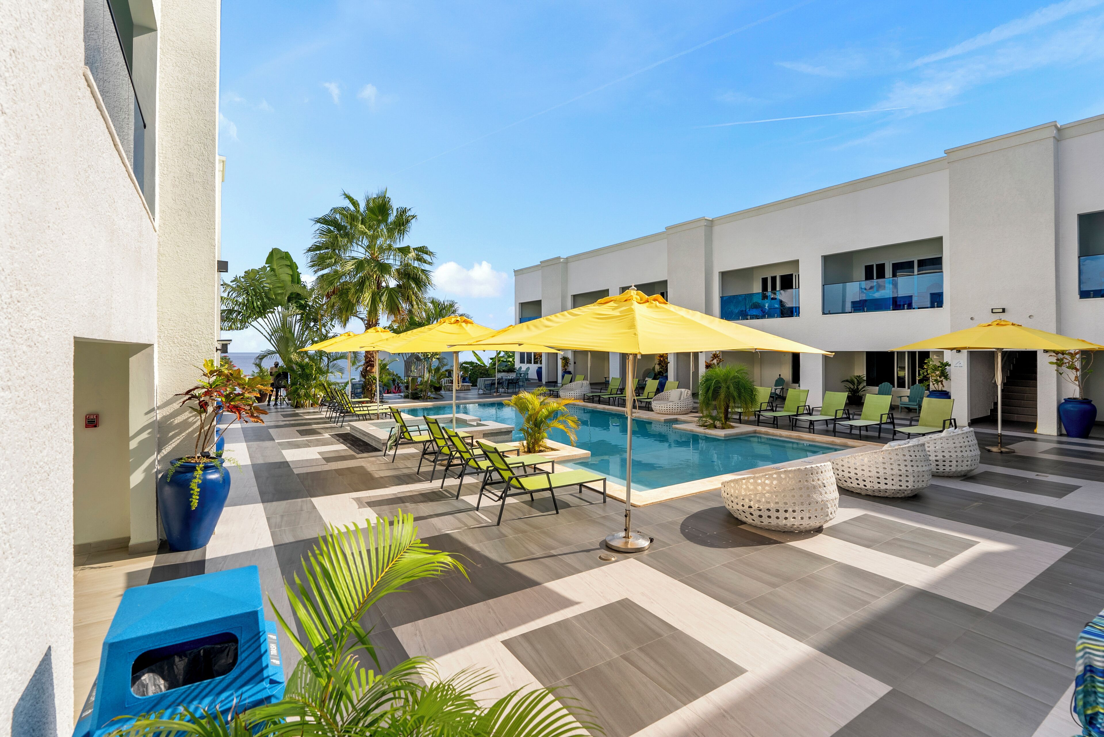 A pool area with sun loungers and umbrellas, surrounded by a white building with multiple windows.