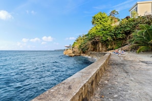 A coastal scene with a concrete walkway along the water, a small structure, and lush greenery.