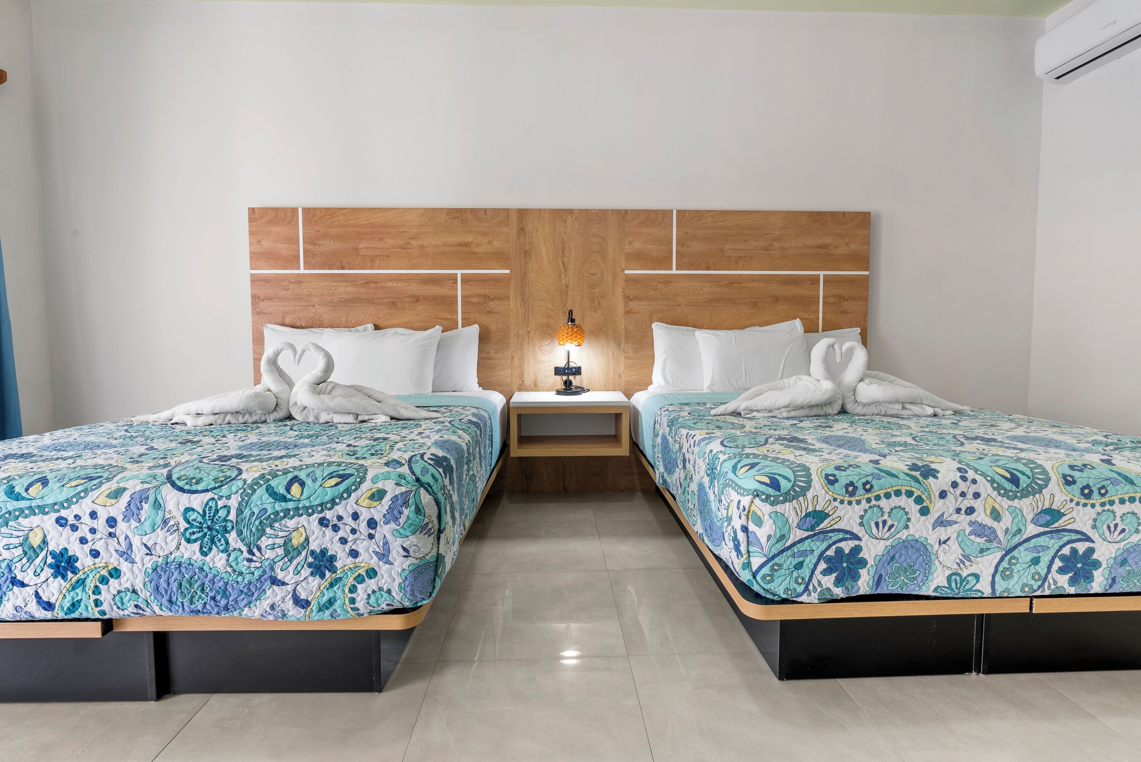 A hotel room with two beds, each with a patterned bedspread, a nightstand with a lamp, and a wooden headboard.