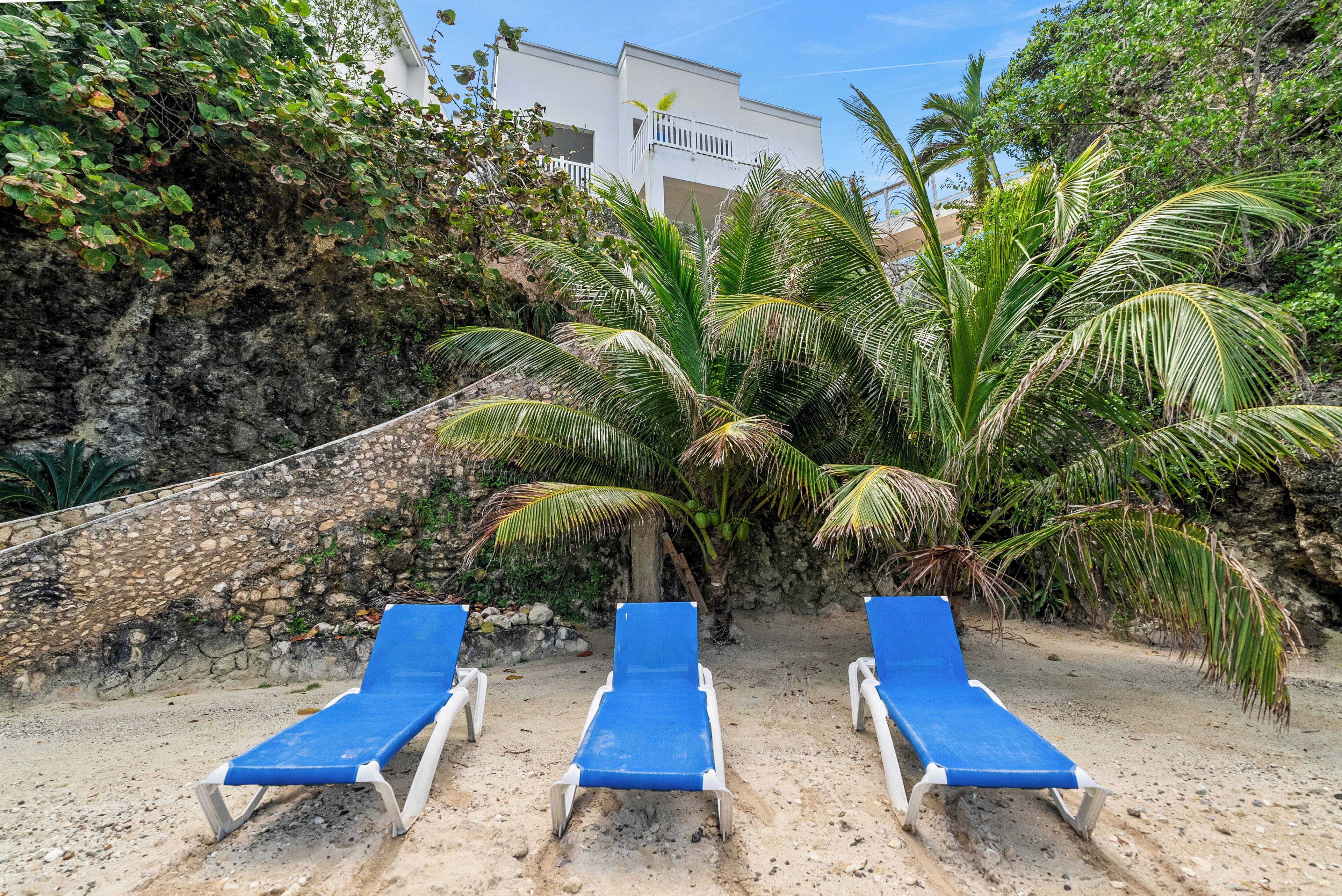 Tres sillones azules sobre una playa arenosa, con un acantilado rocoso y un edificio blanco al fondo.