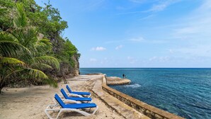 Una playa con sillas azules para tomar el sol, un acantilado rocoso y un mar tranquilo.