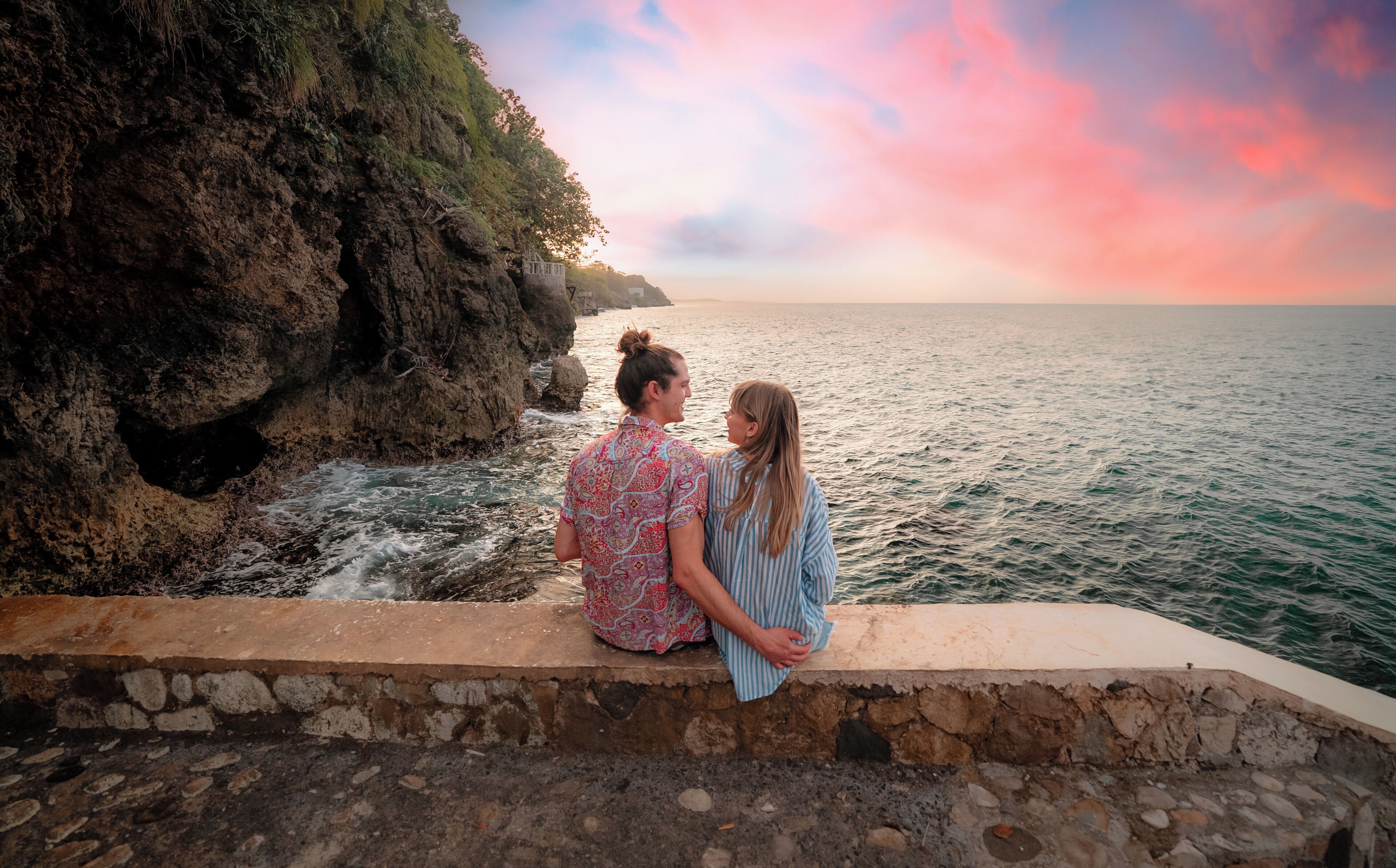 Dos personas sentadas sobre una cornisa de piedra con vista a una costa rocosa y al mar durante el atardecer.