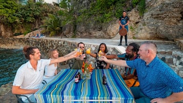 Un grupo de personas brindando con bebidas en una mesa frente a la costa, con un acantilado rocoso y vegetación al fondo.