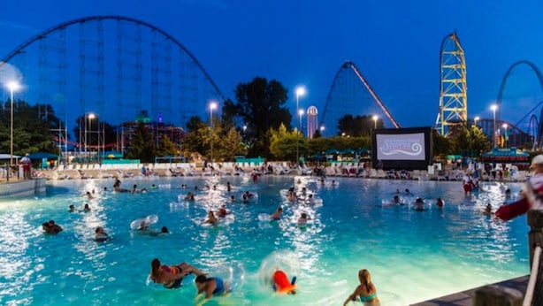 Zona de piscina con gente nadando y jugando, rodeada de atracciones de parque de diversiones y una pantalla grande mostrando un evento.