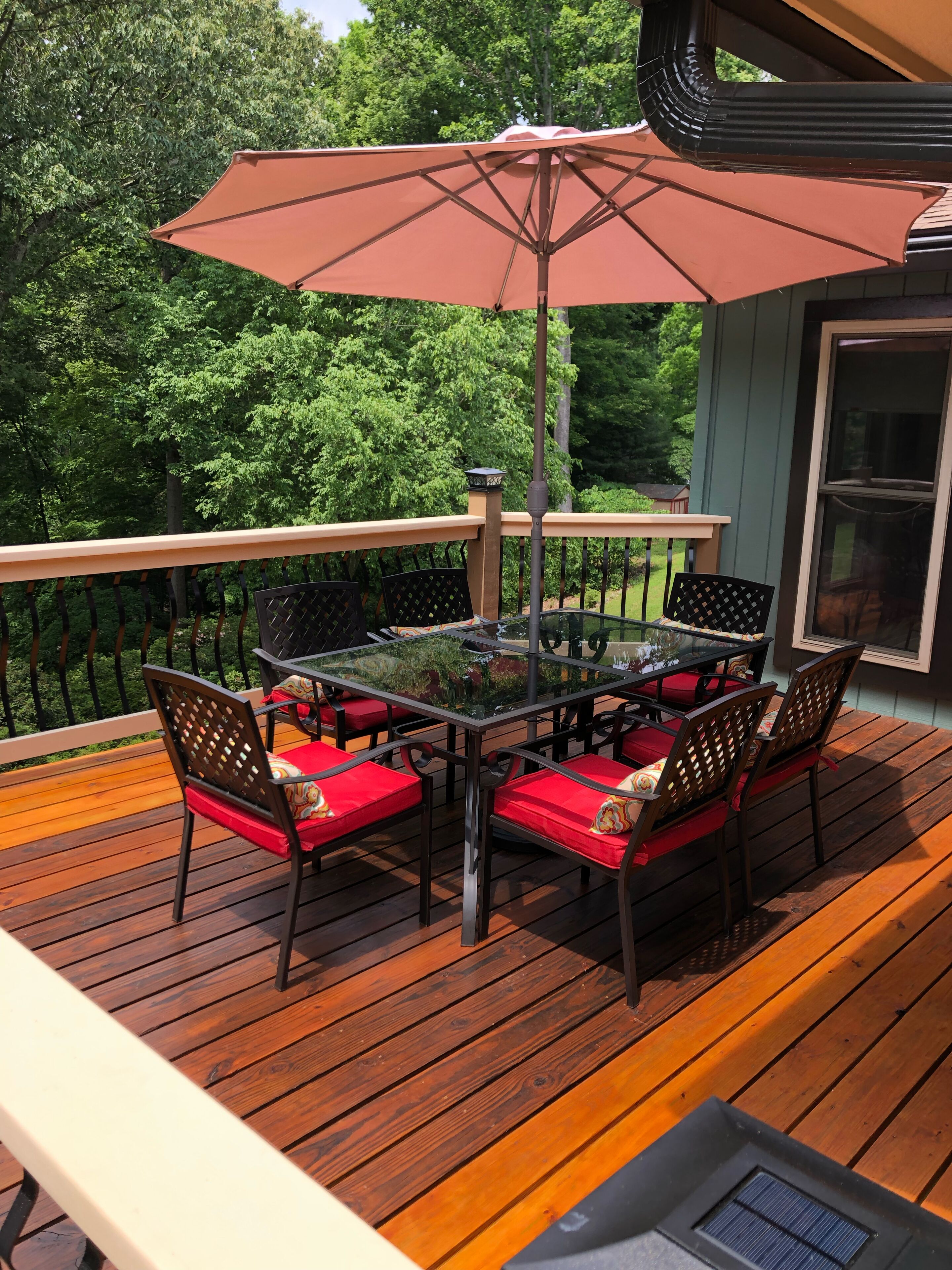 A deck with a glass-top table, red cushioned chairs, and a brown umbrella.
