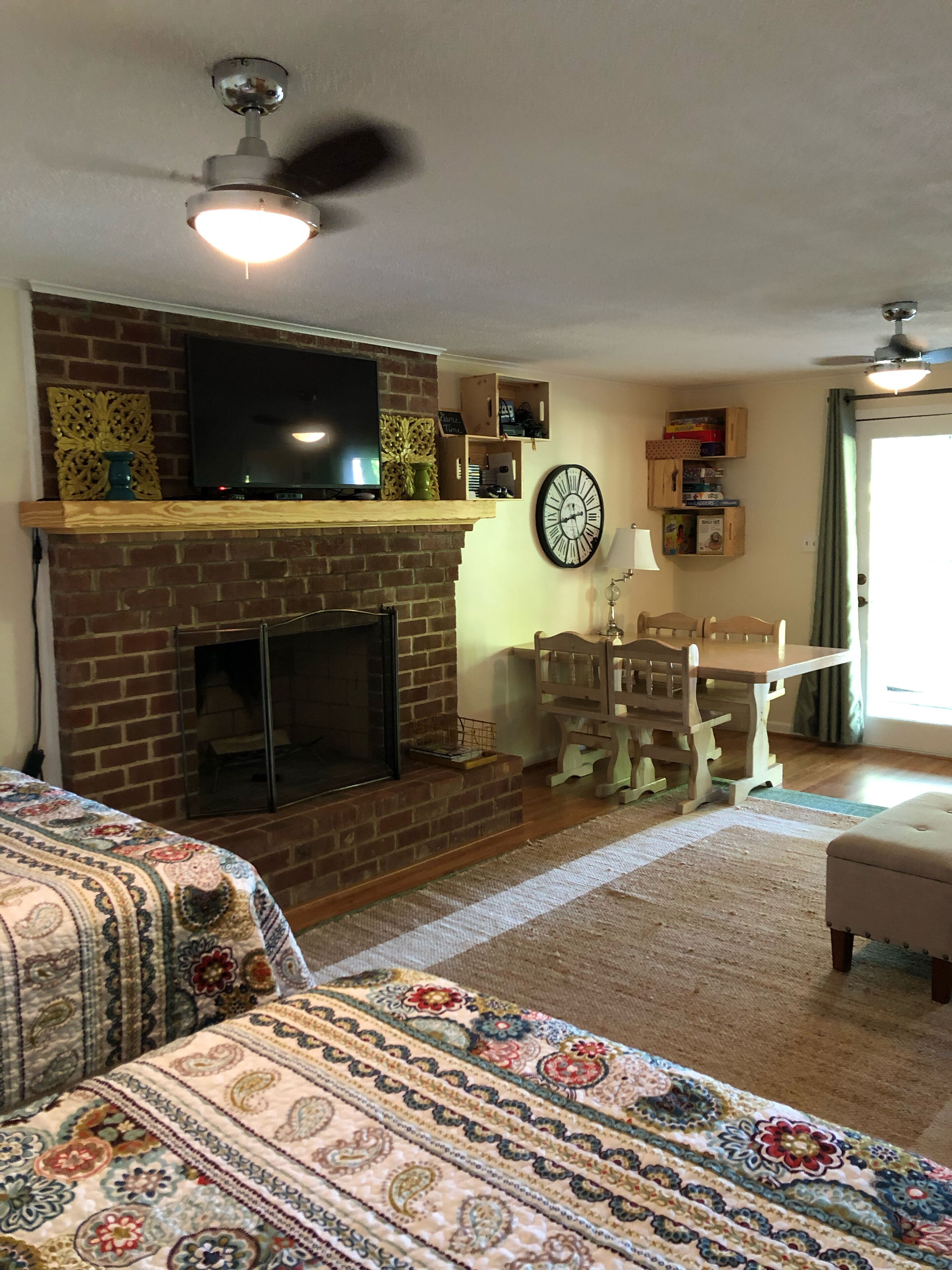 Ein Wohnzimmer mit Kamin aus Backstein, einem Fernseher, einem Esstisch mit Stühlen und einer Wanduhr.