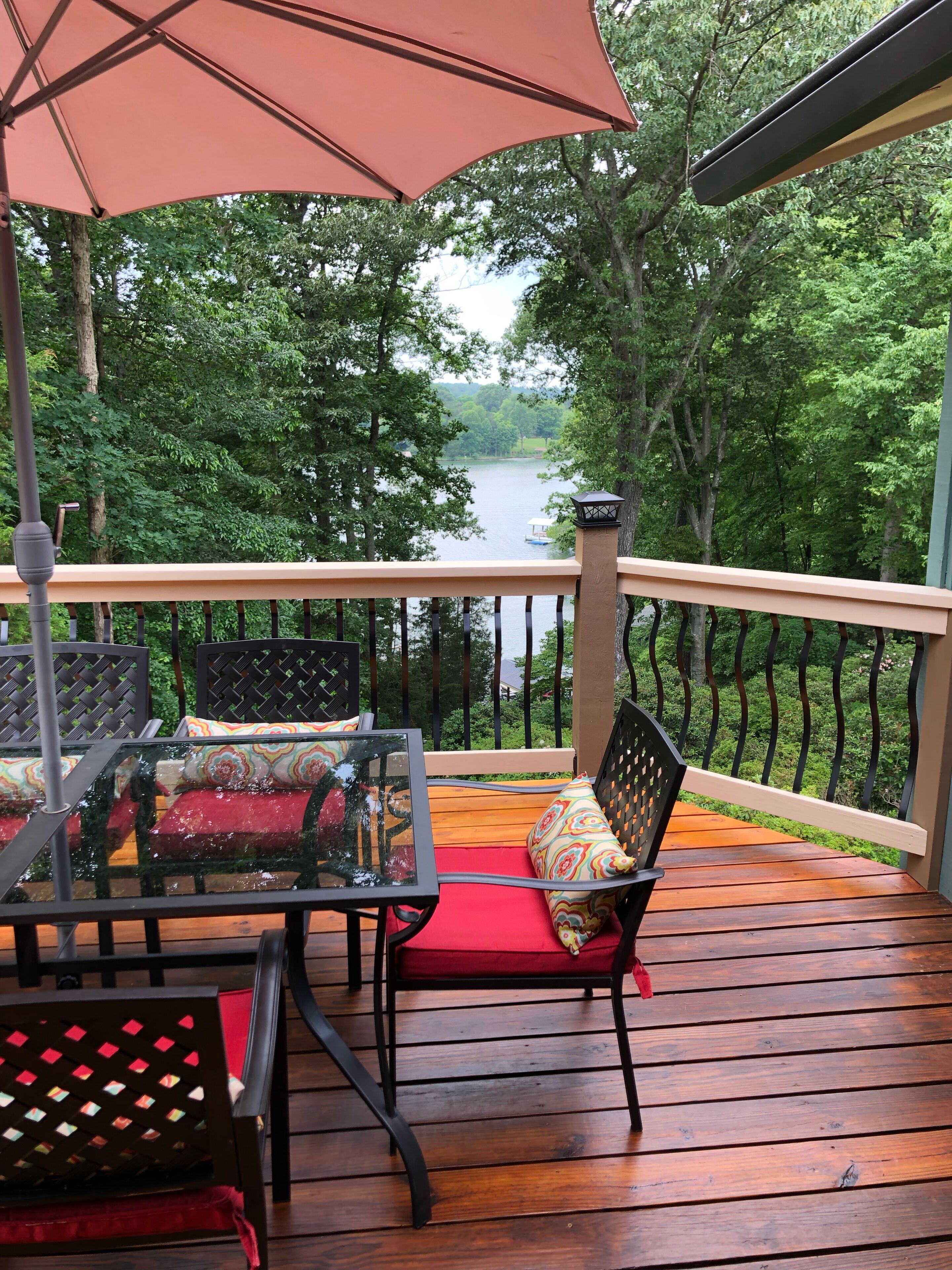 Une terrasse en bois avec une table et des chaises en verre, un banc rouge avec coussin et un grand parasol.