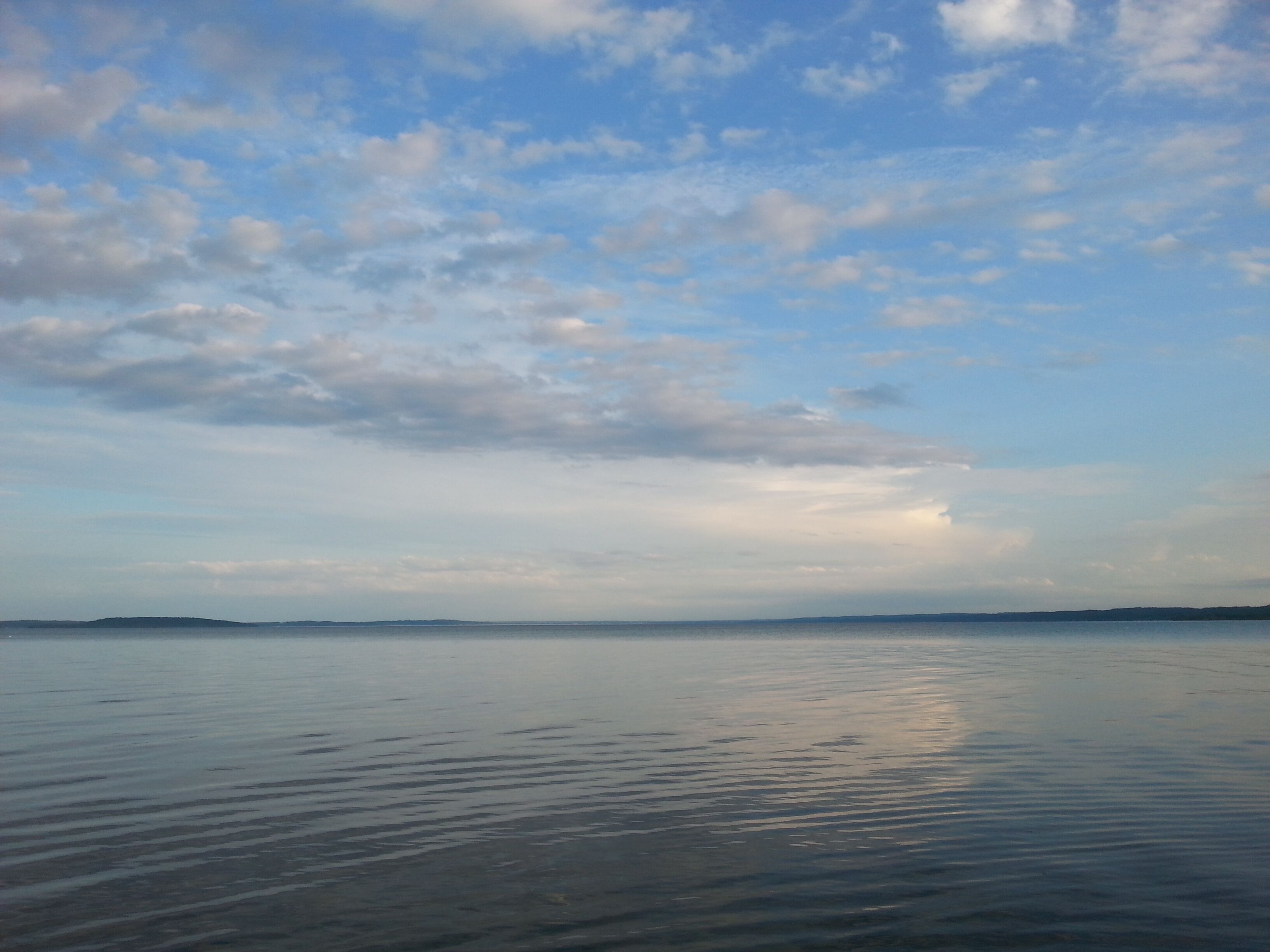 Une étendue d’eau calme, avec une rive lointaine et un ciel partiellement nuageux.