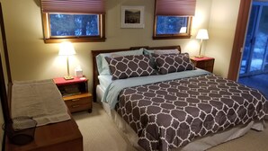 Un dormitorio con cama, dos mesitas de noche con lámparas, una ventana con persianas, un cuadro en la pared y un ventanal con vistas a árboles.