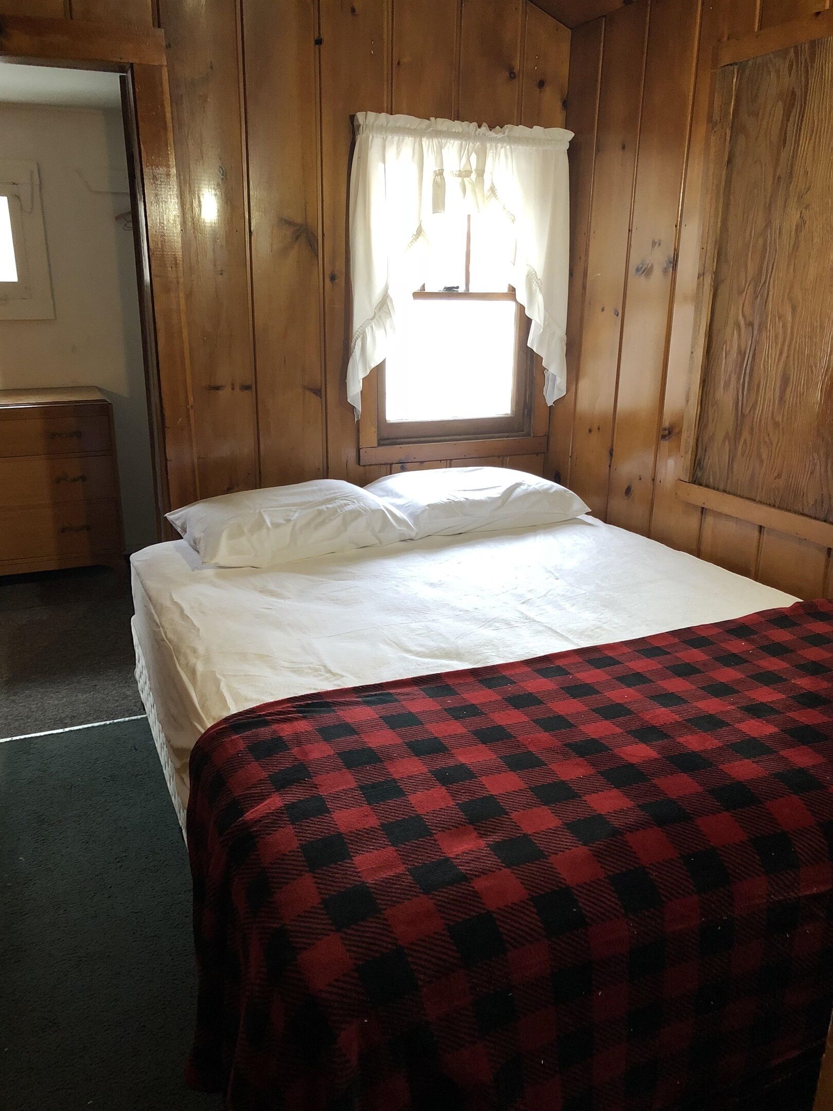 A wooden room with a bed, a window with curtains, and a dresser.