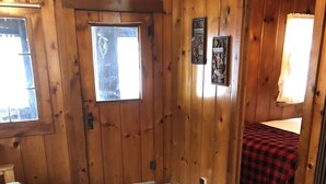 Une pièce aux murs en bois, dotée d’un plancher recouvert de moquette, d’un lit avec une couverture à carreaux rouges et noirs, et de deux fenêtres.