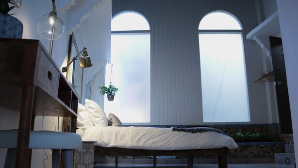 Ein Schlafzimmer mit einem Bett, Nachttischlampen, einer Topfpflanze und großen Bogenfenstern.