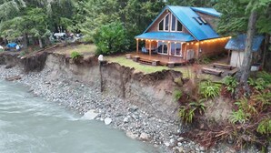 A house with a blue roof and a porch, situated on a cliff by a river. - Relaxing home with an unsettling river bank. (Baring)