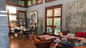 A spacious room with wooden flooring, a dining area with a table and chairs, a sofa with colorful cushions, and large windows with wooden shutters. - BirdHouse@Gamboa, Panama Canal (Gamboa)