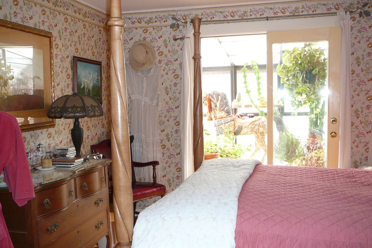 Un dormitorio con una cama con dosel, un aparador, un espejo y una ventana con vistas a zonas verdes.