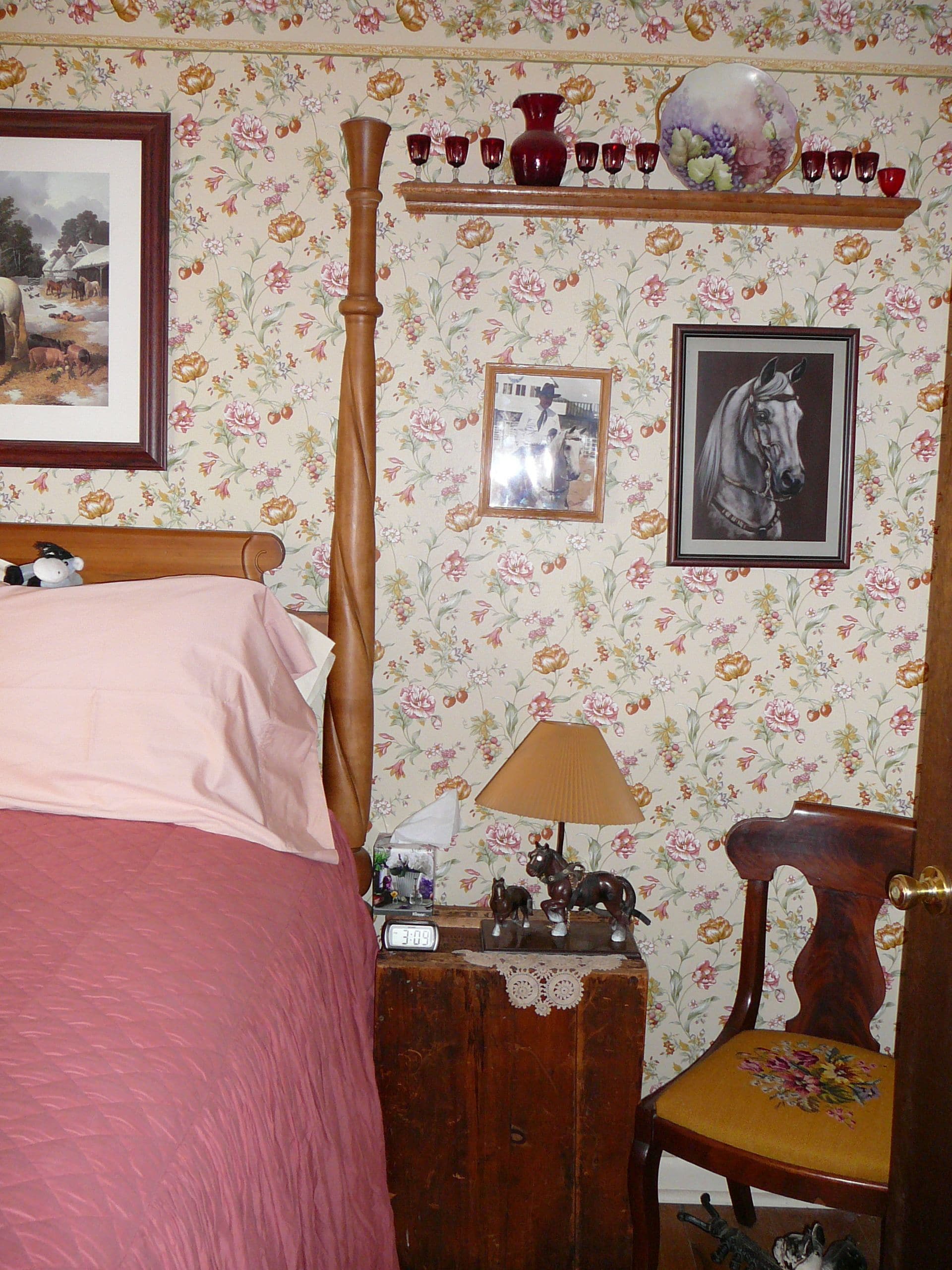 Un dormitorio con cama con dosel, papel tapiz floral, una mesita de noche de madera con una lámpara, una silla con cojín floral y un cuadro enmarcado de un caballo.