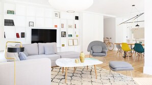 A modern living room with a sofa, coffee table, and armchairs, a flat-screen TV, and a bookshelf with various items.