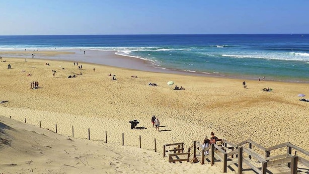 A wide sandy beach with a wooden staircase leading down to the water, and a clear blue sky above.