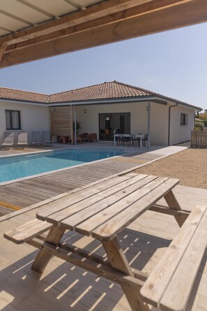 A poolside area with a wooden table and chairs, a covered patio, and a house with a tiled roof.