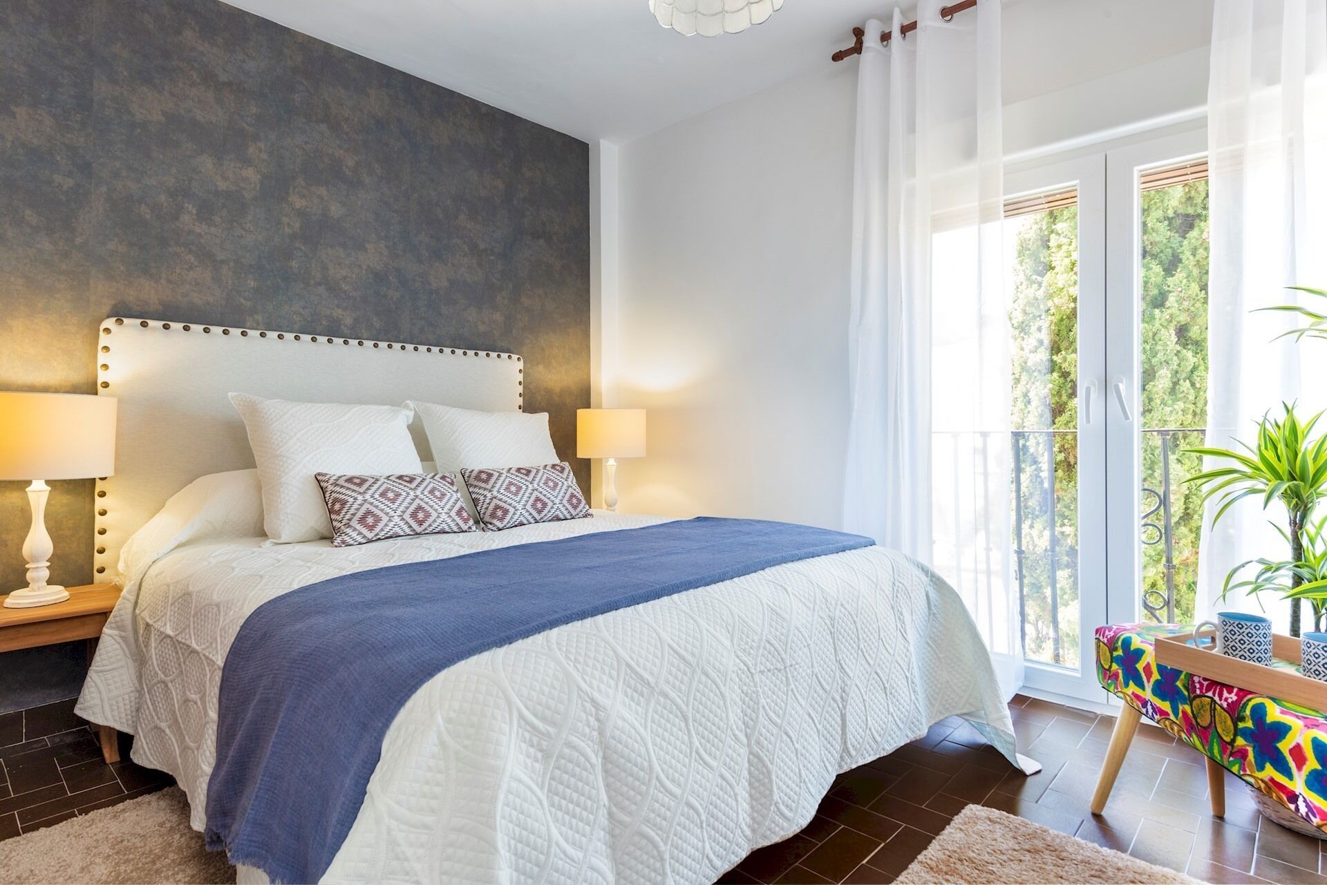 A bedroom with a large bed, two bedside lamps, a blue throw blanket, and a view of greenery through a window.