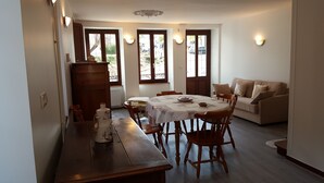 Un comedor con mesa de madera, sillas y un sofá. Se observan ventanas con cortinas, un radiador y luces fijadas a la pared.