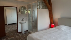 Un dormitorio con cama, lámpara de noche, cabina de ducha de vidrio, lavamanos blanco y armario.
