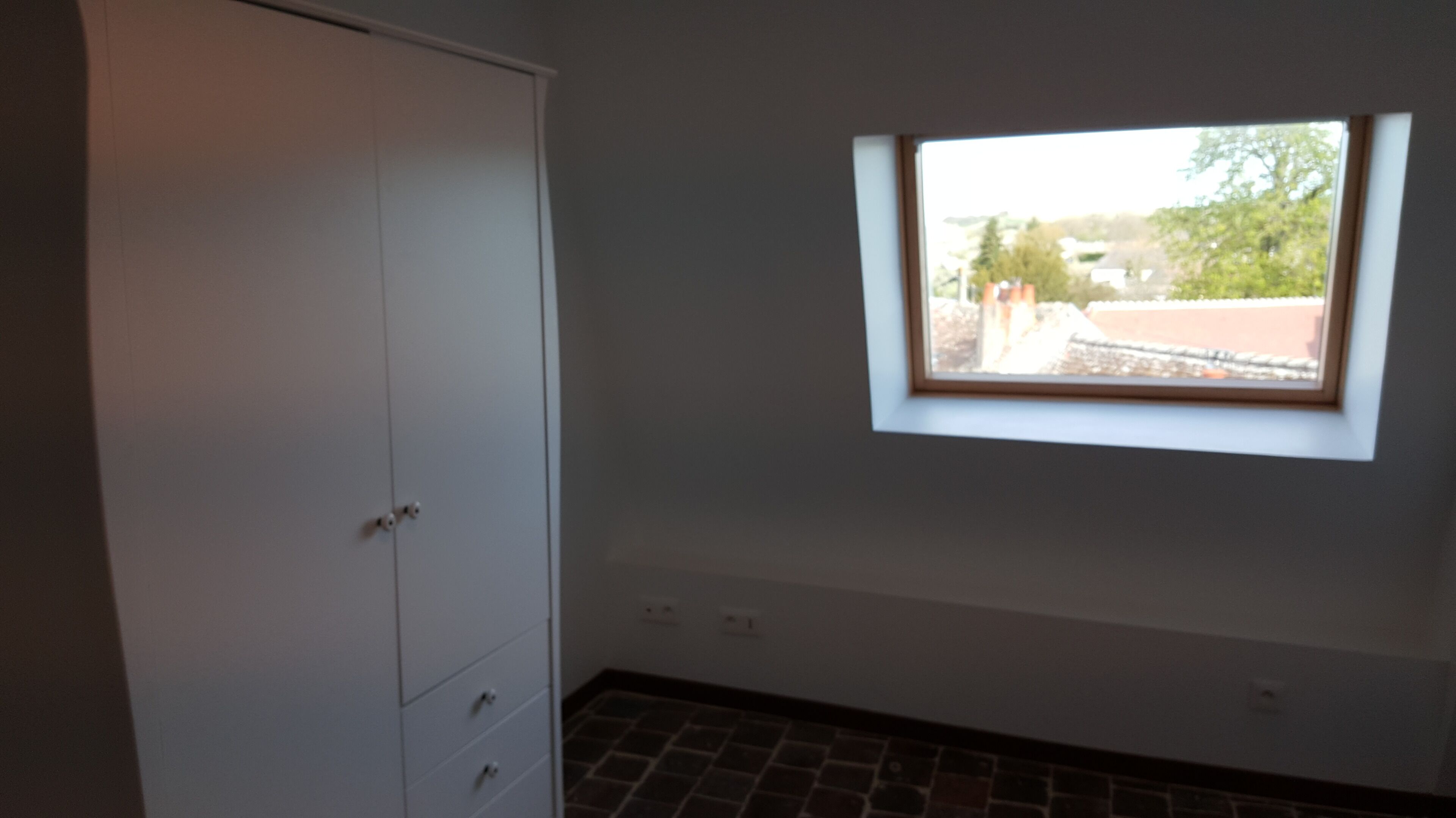 Ein Zimmer mit einem weißen Kleiderschrank, einem kleinen Fenster mit Blick auf Gebäude und Bäume sowie einem Fliesenboden.