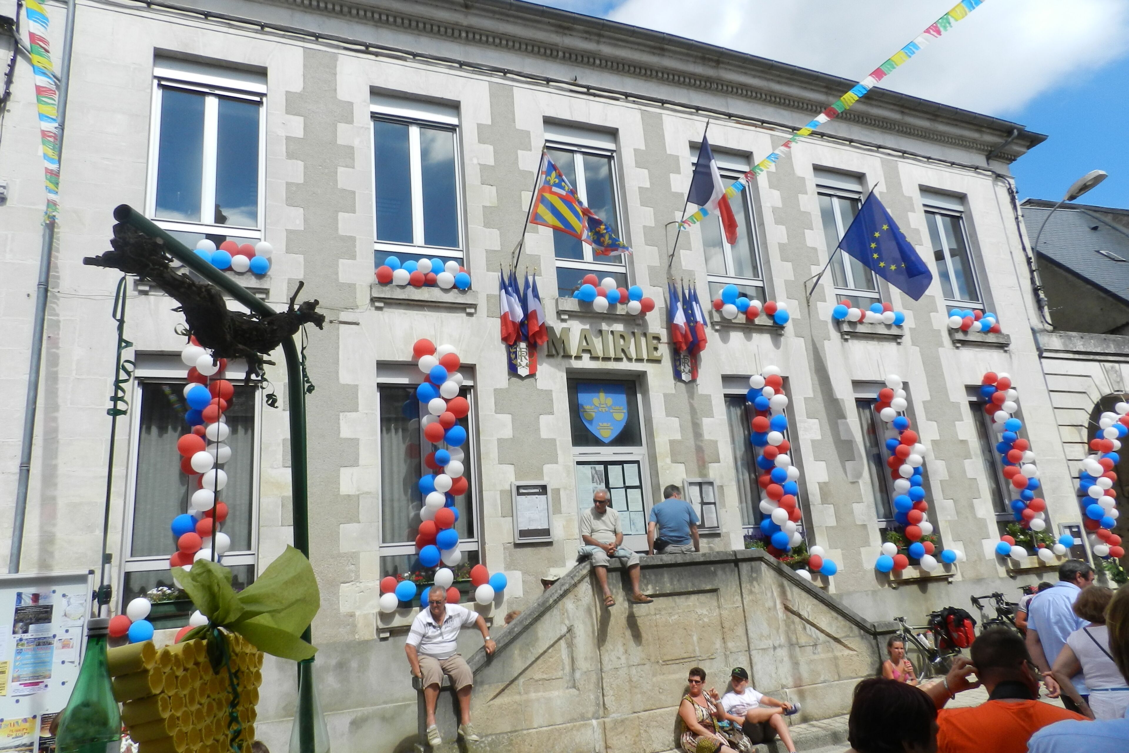 A building with a stone facade, multiple windows, and a sign reading "MAIRIE". Flags and balloons are visible, indicating a festive or official event.