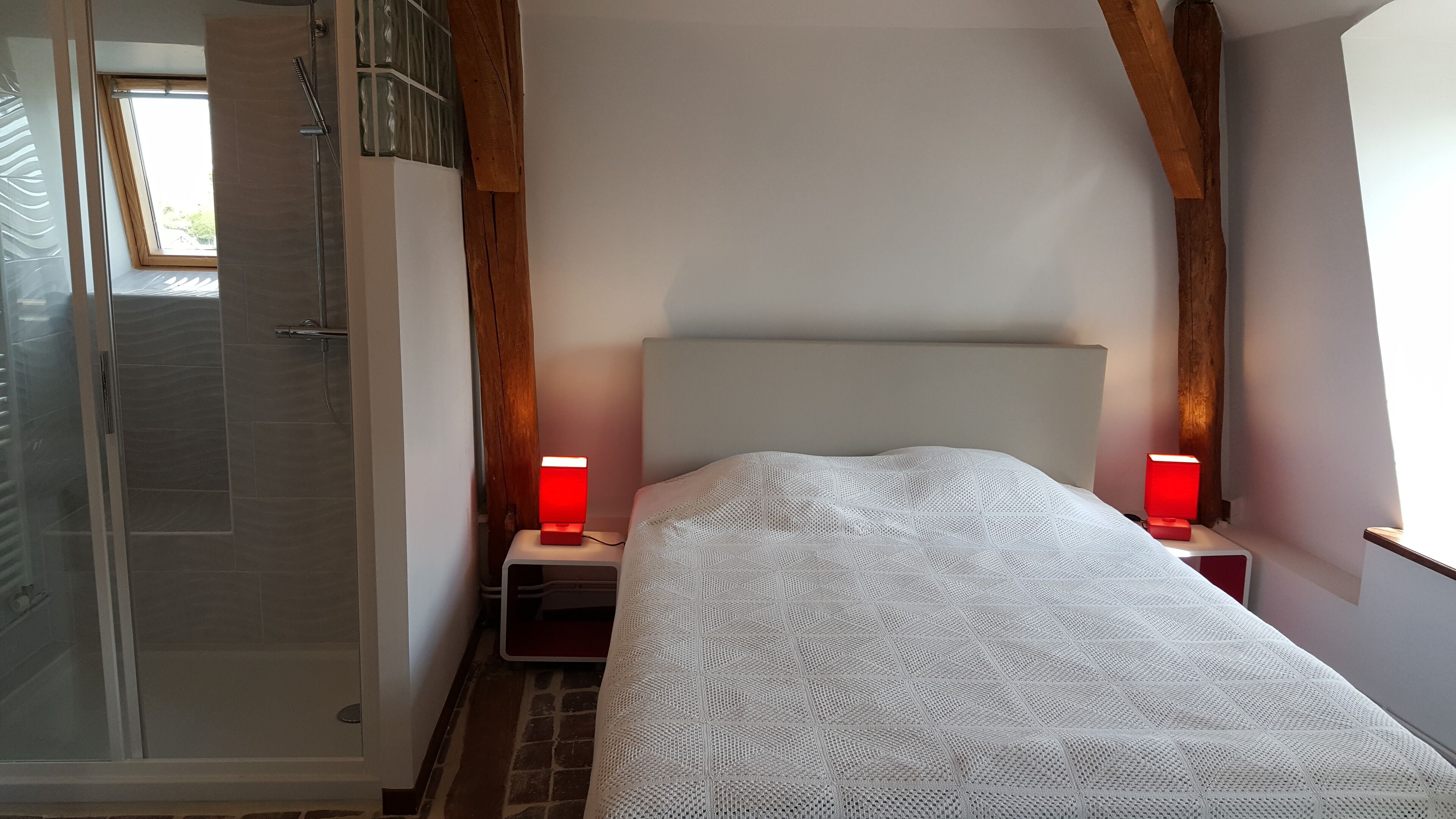 Une chambre à coucher avec un lit, deux tables de chevet dotées de lampes rouges, un plafond en bois et un espace douche.