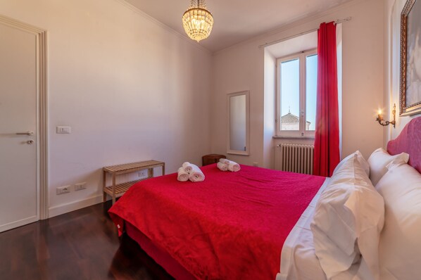 A bedroom with a red bedspread, a wooden bench, a mirror, and a window with red curtains.