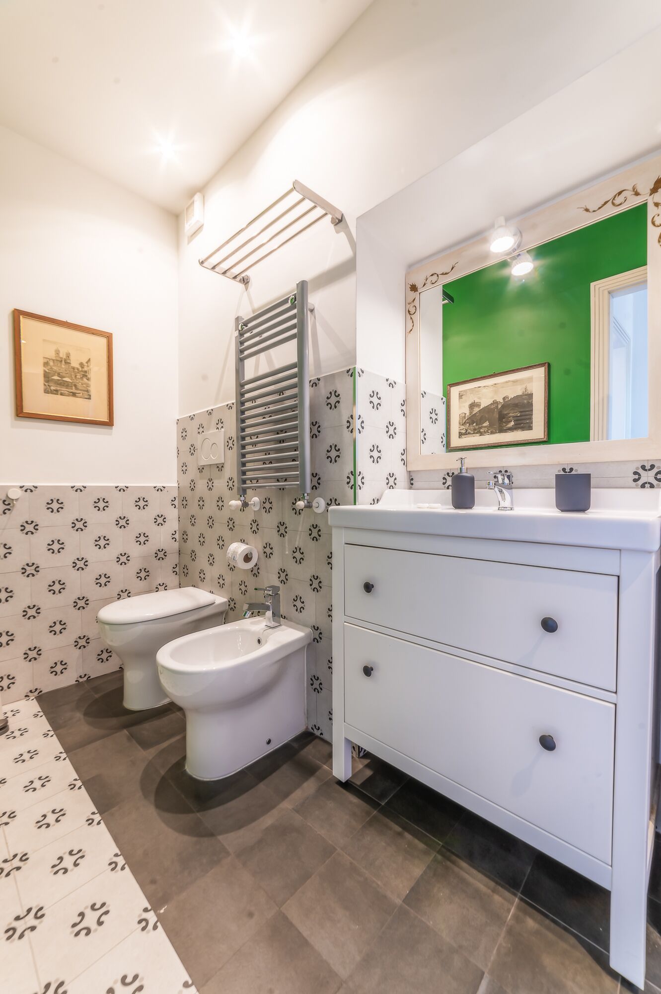 A bathroom with two toilets, a white vanity with drawers, a mirror, and patterned tiles.