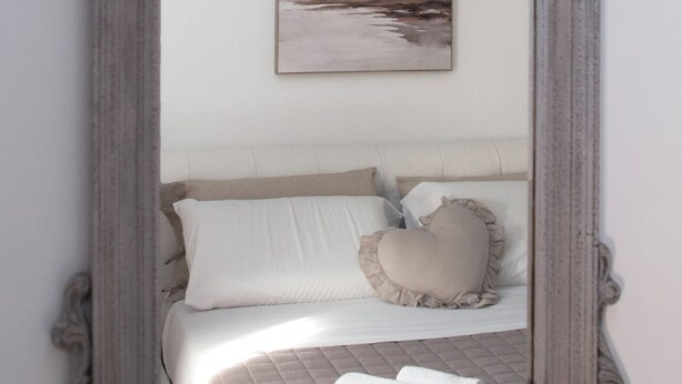 Espejo con marco decorativo que refleja una cama con ropa de cama blanca y un almohadón.