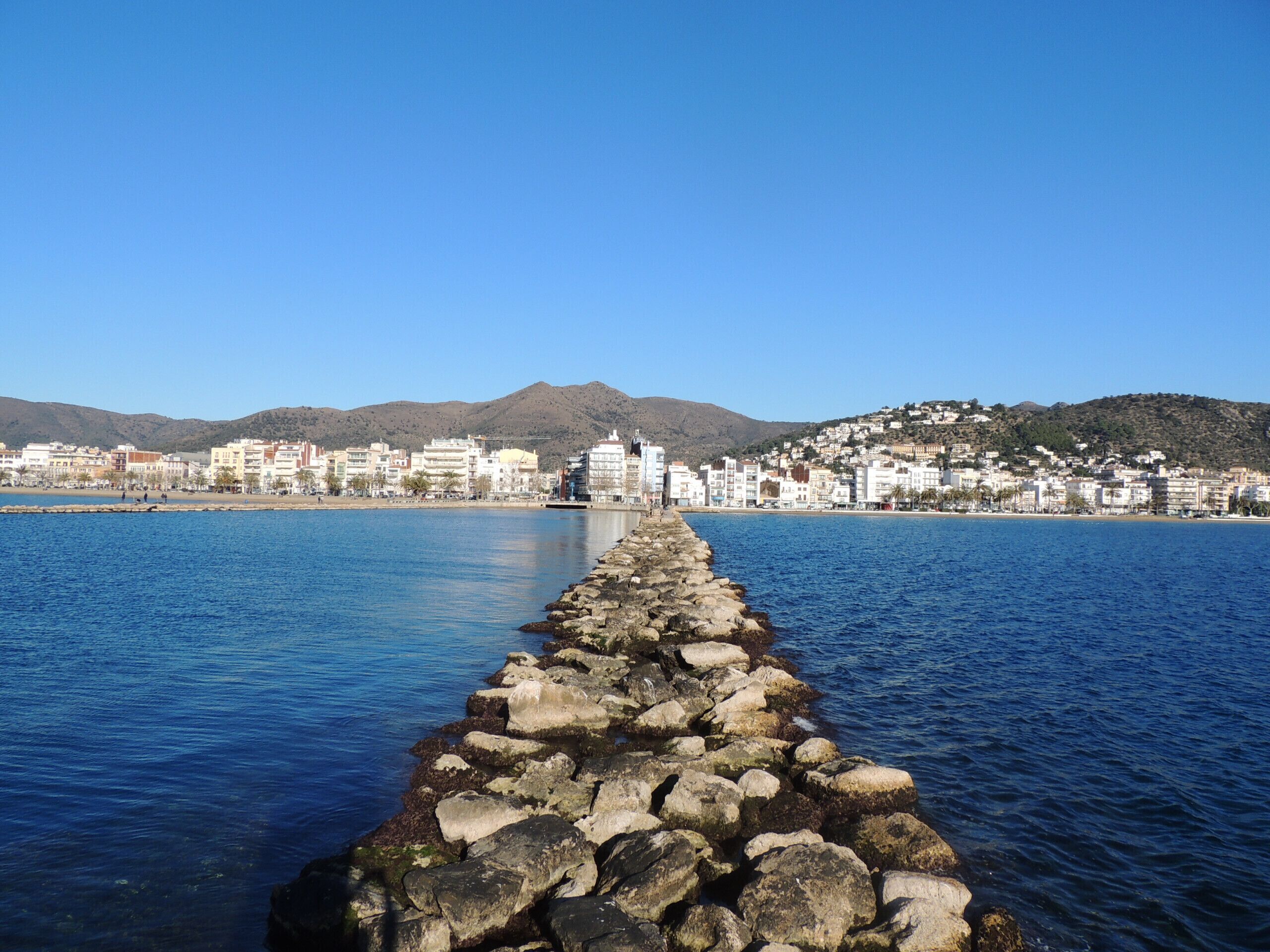 Paesaggio costiero con un molo roccioso che si protende verso il mare, edifici sullo sfondo e una catena montuosa.