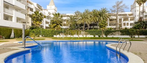 A swimming pool surrounded by a landscaped area with palm trees and a building with a pointed roof in the background.
