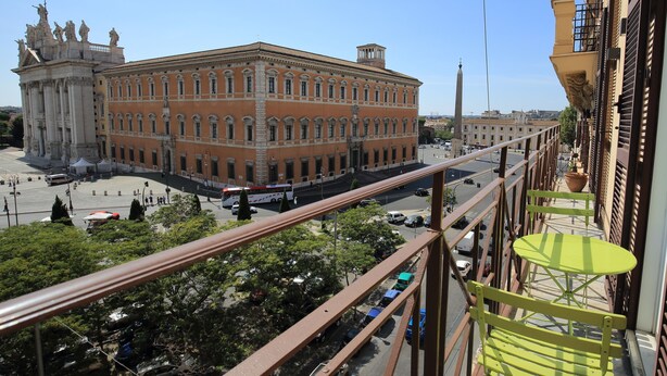 Vista desde un balcón sobre una calle concurrida, con un edificio destacado de arquitectura clásica y estatuas.