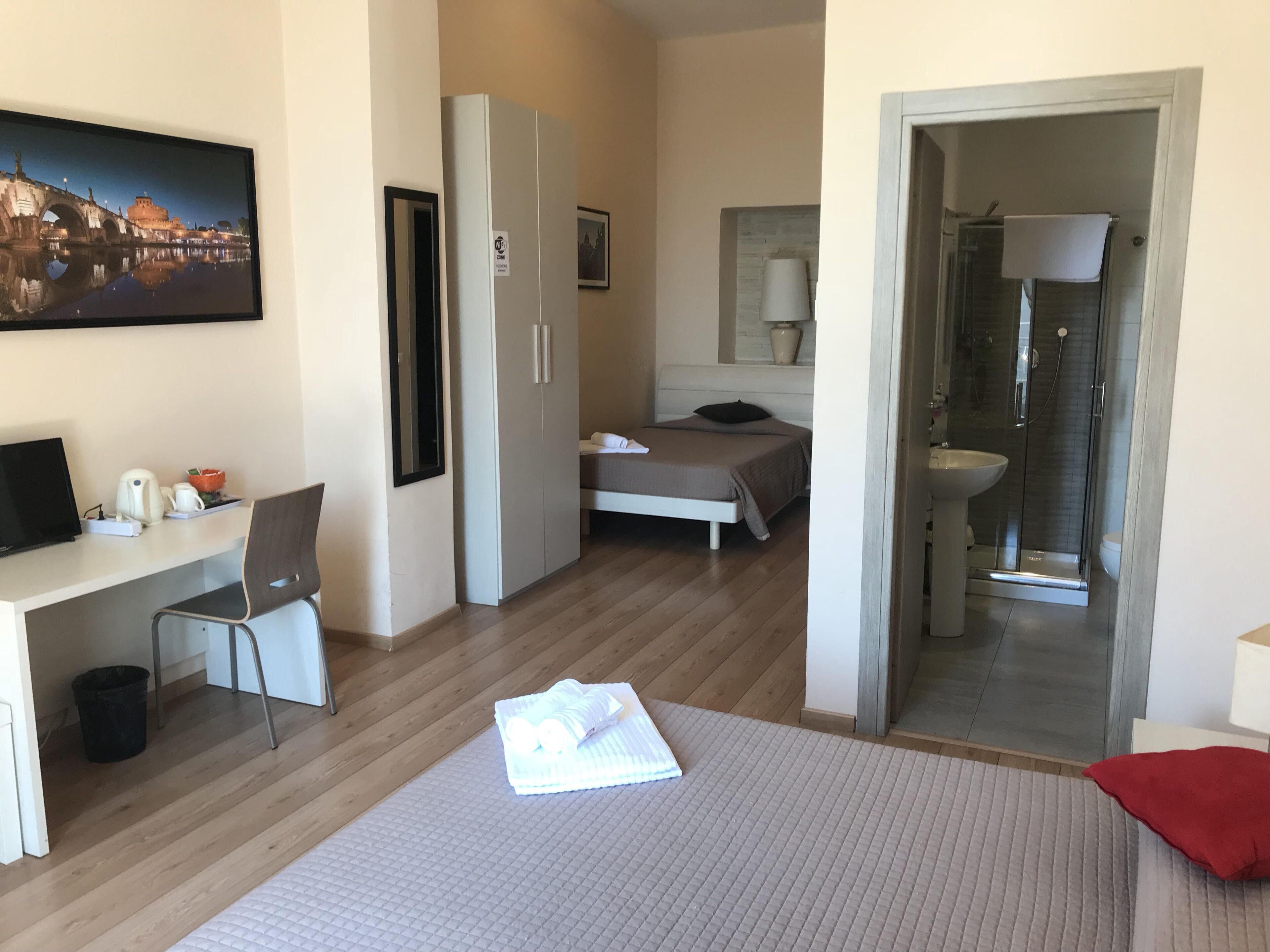A hotel room with a bed, a desk, a chair, a wardrobe, a bathroom with a shower, and a painting of a cityscape.
