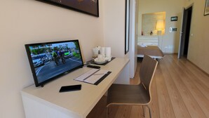 Habitación de hotel con escritorio, televisión mostrando una prueba de ciclismo, una silla y una cama al fondo.
