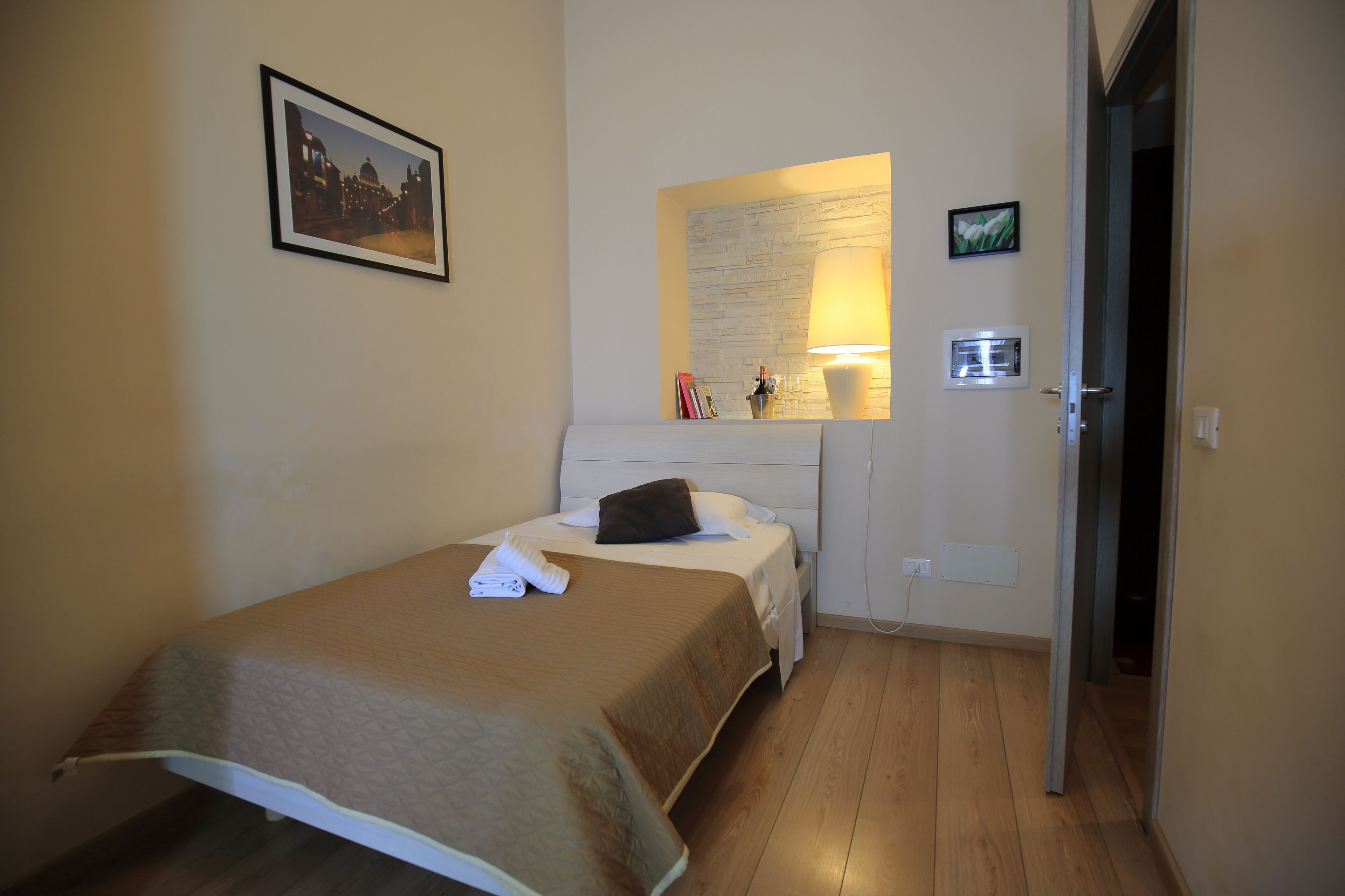 A small, neatly arranged bedroom with a bed, bedside table, lamp, and a framed picture on the wall.