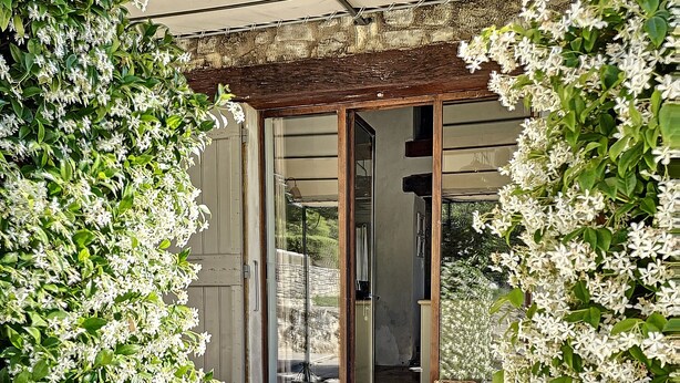 Un espace extérieur abrité, doté d’une porte donnant sur une pièce, et entouré d’une végétation luxuriante et de fleurs blanches.