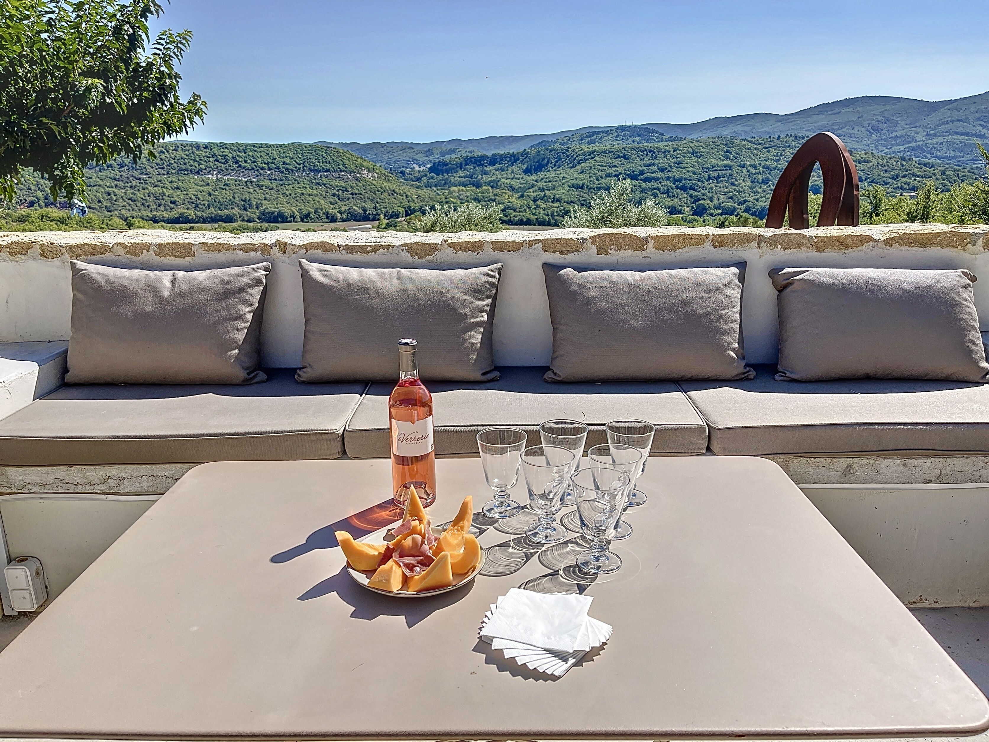 A terrace with a table set for a meal, a bottle of wine, and glasses. The terrace overlooks a scenic landscape with hills and trees.