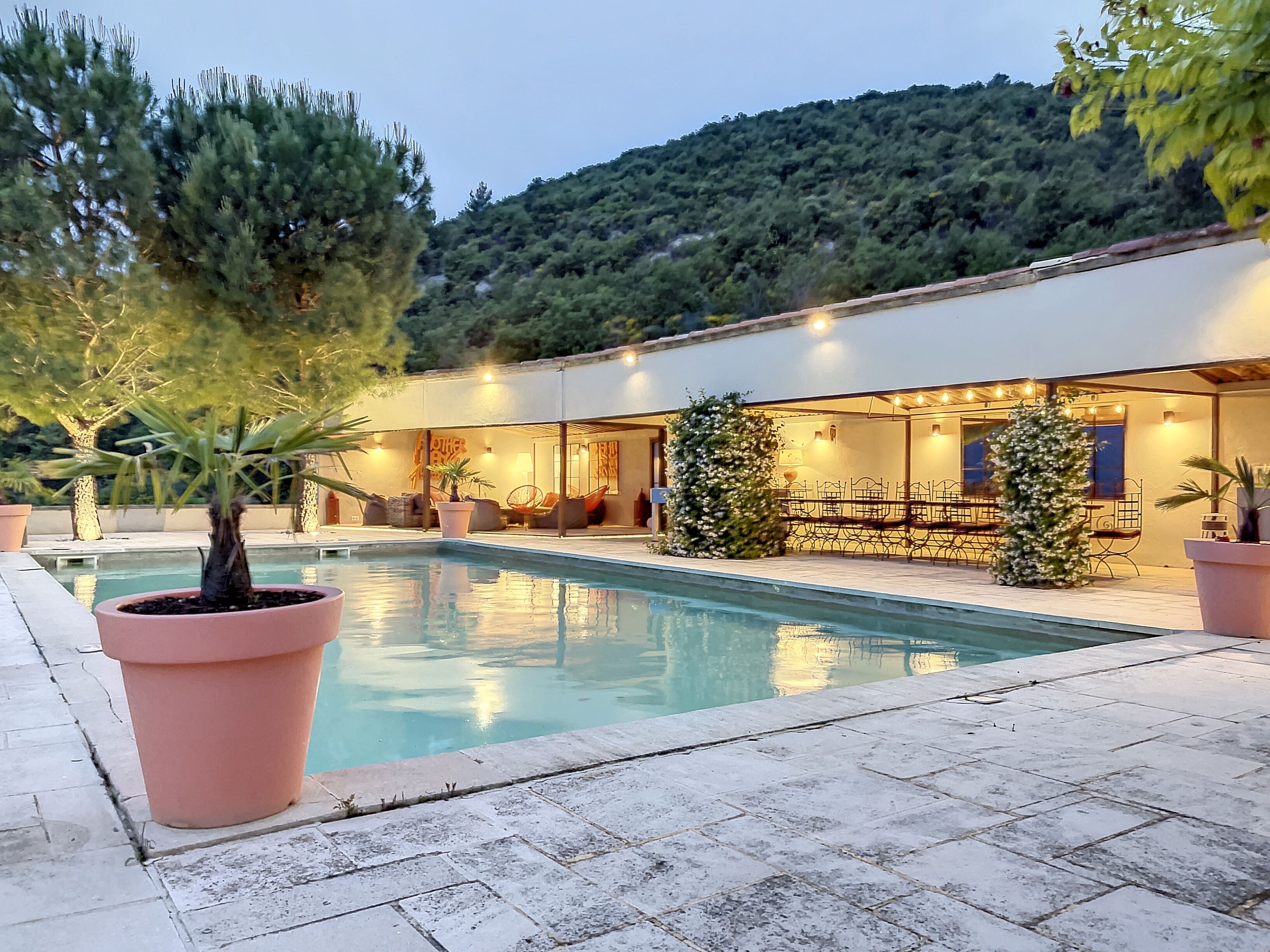Un espace piscine avec une terrasse, des plantes en pot et un bâtiment doté d’un espace extérieur aménagé pour s’asseoir.