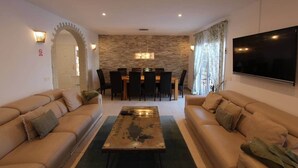 A living room with two sofas, a coffee table, a dining table with chairs, a stone wall, and a TV mounted on the wall. - ViVa Tropical (Buzanada, Arona)