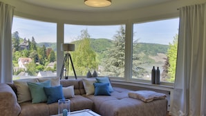 A living room with a large sectional sofa, a glass coffee table, and a floor lamp. The room has large windows offering a view of a green landscape.