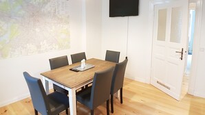 A dining area with a wooden table, six chairs, a map on the wall, a TV mounted above the table, and a door with frosted glass panels.