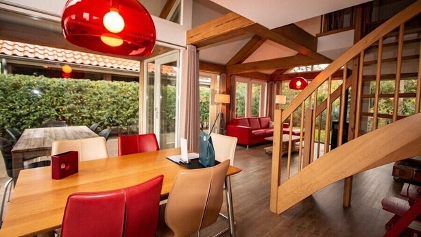 Un comedor con una mesa de madera y sillas rojas, una escalera con barandillas de madera y vista a zonas verdes exteriores.