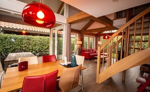 A dining area with a wooden table and red chairs, a staircase with wooden railings, and a view of greenery outside.