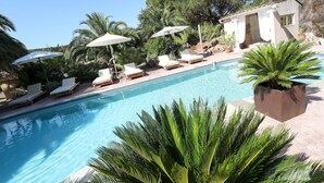 A pool area with lounge chairs and umbrellas, surrounded by greenery and a building with a stone facade.