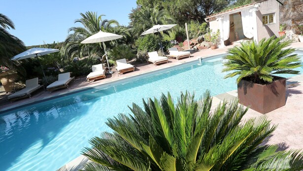 A pool area with lounge chairs and umbrellas, surrounded by greenery and a building with a stone facade.