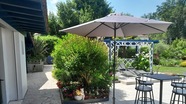 Un patio con mesa y sillas, una sombrilla grande y un jardín con diversas plantas y flores.