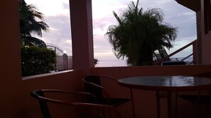 A balcony with a round table and two chairs, overlooking a palm tree and the ocean.